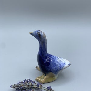 May include: A small ceramic goose figurine with blue and white glaze. The goose is standing on a light gray surface with lavender flowers.