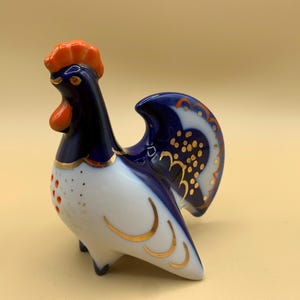 May include: A blue and white porcelain rooster figurine with gold accents. The rooster has an orange comb and wattle and a blue tail.