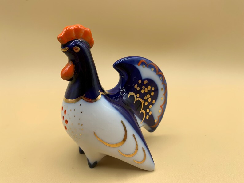 May include: A blue and white porcelain rooster figurine with gold accents. The rooster has an orange comb and wattle and a blue tail.