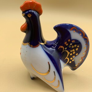 May include: A porcelain rooster figurine with blue and white paint and gold accents. The rooster has a red comb and wattle.