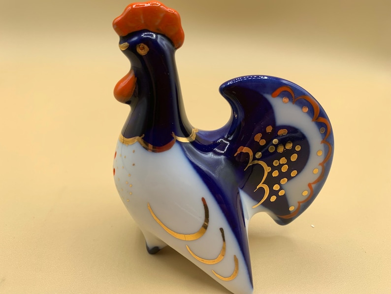 May include: A porcelain rooster figurine with blue and white paint and gold accents. The rooster has a red comb and wattle.