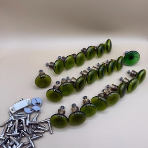 May include: A collection of green glass cabinet knobs with metal hardware. The knobs are arranged in rows, with a variety of screws and mounting hardware. The knobs have a rounded, oval shape and a vintage aesthetic.