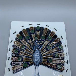 May include: A white ceramic tile with a raised design of a peacock with its tail feathers spread out. The peacock is blue, green, and brown. The tile is signed "Rörstrand Sweden".