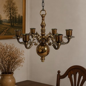 Può includere: Un lampadario in ottone lucido con sei portacandele disposti attorno a un corpo centrale sferico. L'apparecchio è sospeso da una catena in metallo scuro e presenta bracci curvi decorati con volute dettagliate che collegano ogni piattino.
