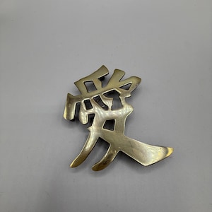 May include: A brass-coloured metal pin in the shape of a Chinese character, 愛, meaning "love." The pin has a slightly aged appearance and is set against a grey background. The character's design is detailed and three-dimensional.