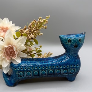 May include: A blue ceramic cat-shaped vase with a textured finish. The vase contains a bouquet of artificial flowers in white, pink, and beige. The cat has a detailed face and geometric patterns along its body.