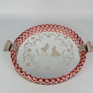 Op de afbeelding: Een rond dienblad met spiegel en een gedraaide glazen rand in rood en transparant. De centrale spiegel toont een geëtst ontwerp van twee figuren in historische kleding omringd door bloemmotieven, met zilverkleurige metalen handgrepen.