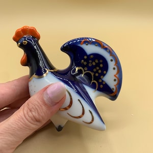 May include: A blue and white porcelain rooster figurine with gold accents. The rooster has a red comb and wattle and a blue tail.