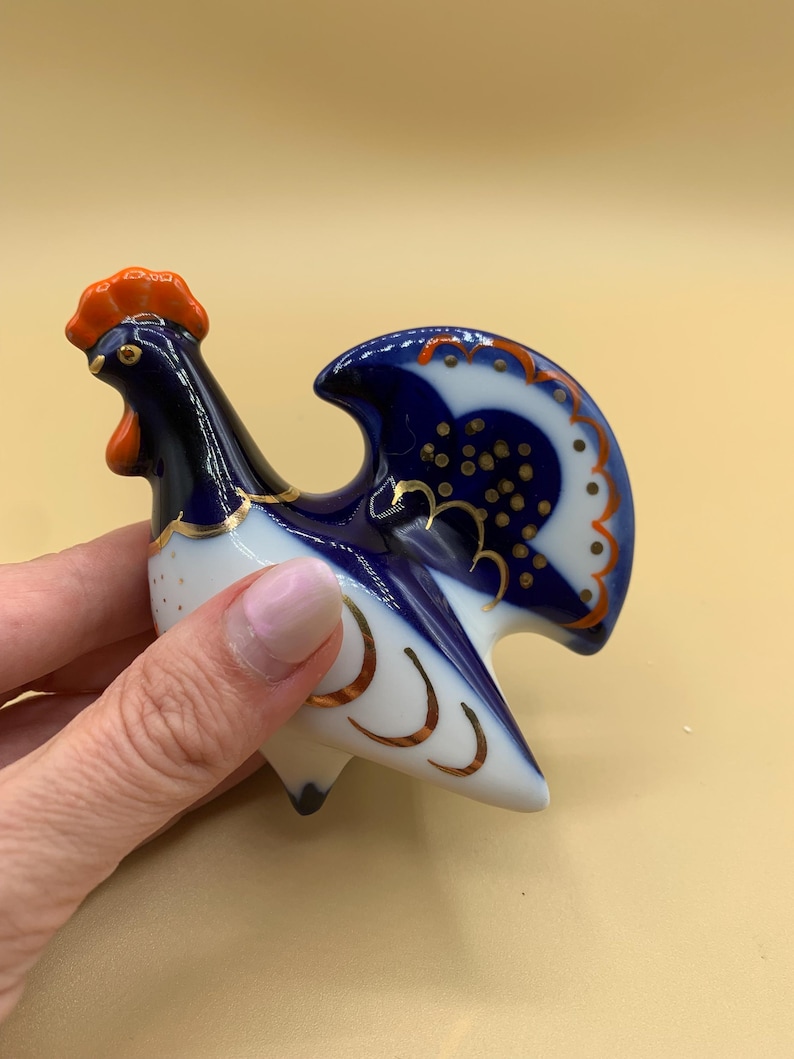 May include: A blue and white porcelain rooster figurine with gold accents. The rooster has a red comb and wattle and a blue tail.