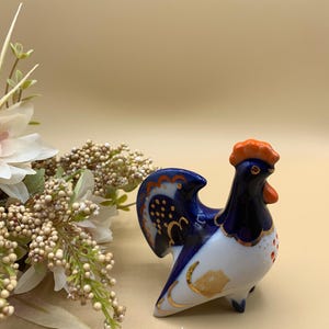 May include: A blue and white porcelain rooster figurine with orange accents. The rooster is standing on a beige surface.