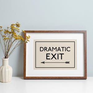 May include: A framed print with the words "DRAMATIC EXIT" in black, with an arrow pointing left. The frame is brown, and the print is on a cream-coloured background. A vase with yellow flowers is to the left.