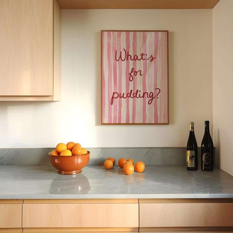 Op de afbeelding: Een keukeninterieur met een ingelijst kunstwerk met de tekst "What's for pudding?" boven een aanrecht. Een terracotta kom gevuld met sinaasappels staat naast losse sinaasappels. Twee wijnflessen staan op het aanrecht.