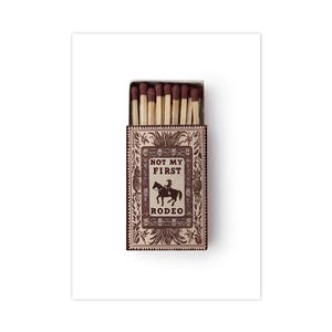 May include: A rectangular matchbox with a brown and cream decorative border featuring a central illustration of a person riding a horse. The text NOT MY FIRST RODEO is printed in the center. The box is open, revealing several wooden matches.