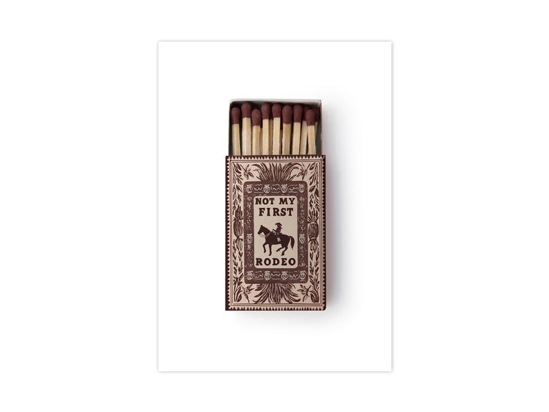 May include: A rectangular matchbox with a brown and cream decorative border featuring a central illustration of a person riding a horse. The text NOT MY FIRST RODEO is printed in the center. The box is open, revealing several wooden matches.