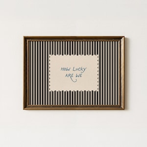 K&ouml;nnte beinhalten: Gerahmtes Wandbild mit schwarz-wei&szlig; gestreiftem Hintergrund. Das Kunstwerk zeigt den handgeschriebenen Text "How Lucky Are We" in Blau auf einem cremefarbenen, gezackten Rechteck. Der Rahmen ist dunkelbraun.
