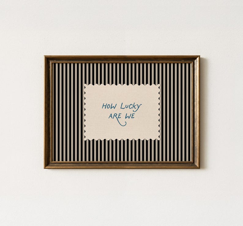 K&ouml;nnte beinhalten: Gerahmtes Wandbild mit schwarz-wei&szlig; gestreiftem Hintergrund. Das Kunstwerk zeigt den handgeschriebenen Text "How Lucky Are We" in Blau auf einem cremefarbenen, gezackten Rechteck. Der Rahmen ist dunkelbraun.