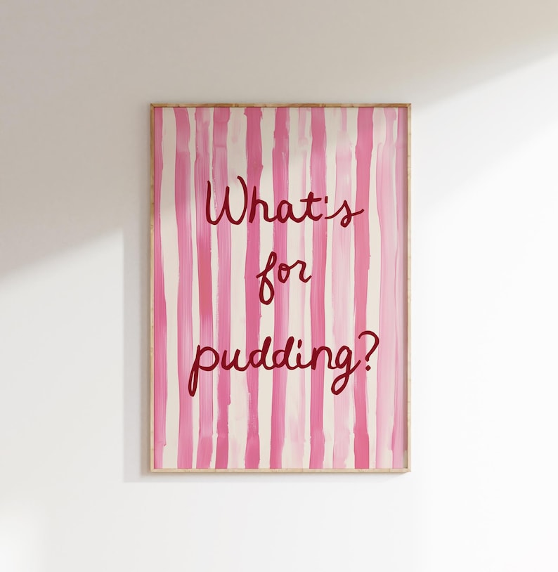 Op de afbeelding: Een ingelijste kunstafdruk met een roze en witte gestreepte achtergrond. De tekst "What's for pudding?" is geschreven in een donkerroze script. Het kunstwerk wordt op een witte muur getoond.
