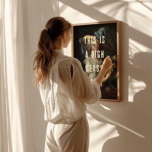 K&ouml;nnte beinhalten: Ein gerahmter Kunstdruck mit einem klassischen Portr&auml;t und dem Text "THIS IS A HIGH CLASS" in Wei&szlig;. Das Kunstwerk befindet sich in einem Holzrahmen. Eine Person in einem wei&szlig;en Hemd und einer Hose h&auml;lt den gerahmten Druck.