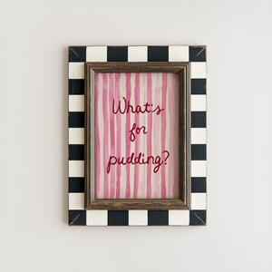 Op de afbeelding: Een ingelijste print met de tekst "What's for pudding?" in rood script. De print heeft roze verticale strepen. De lijst heeft een zwart-wit geblokt patroon aan de buitenrand en een bruine binnenlijst.