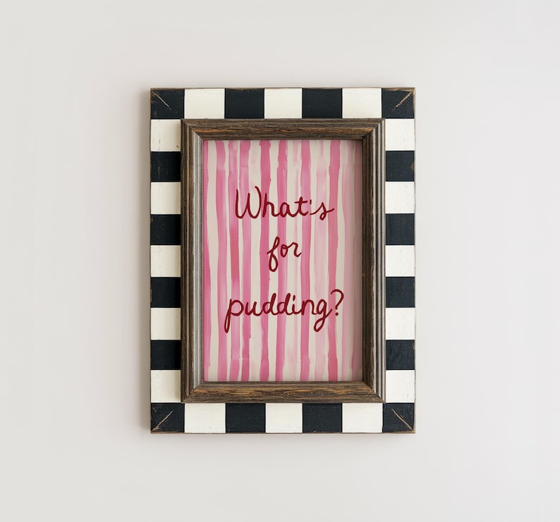 Op de afbeelding: Een ingelijste print met de tekst "What's for pudding?" in rood script. De print heeft roze verticale strepen. De lijst heeft een zwart-wit geblokt patroon aan de buitenrand en een bruine binnenlijst.