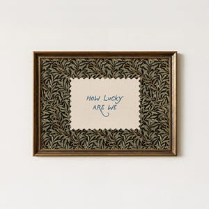 K&ouml;nnte beinhalten: Ein gerahmter Kunstdruck mit einem goldfarbenen Rahmen. Der Druck zeigt ein cremefarbenes, gezacktes Rechteck mit dem handgeschriebenen Text "How Lucky ARE WE" in Blau. Der Hintergrund des Drucks ist ein dunkles Muster aus Bl&auml;ttern.