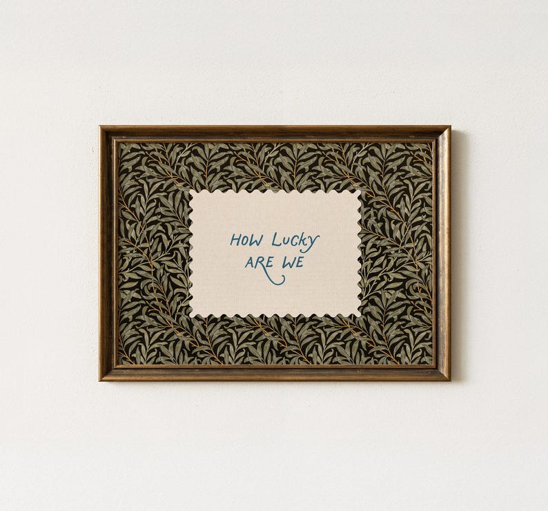 K&ouml;nnte beinhalten: Ein gerahmter Kunstdruck mit einem goldfarbenen Rahmen. Der Druck zeigt ein cremefarbenes, gezacktes Rechteck mit dem handgeschriebenen Text "How Lucky ARE WE" in Blau. Der Hintergrund des Drucks ist ein dunkles Muster aus Bl&auml;ttern.