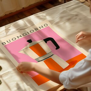Puede incluir: Un p&oacute;ster con una ilustraci&oacute;n estilizada de una cafetera italiana en naranja, blanco y negro sobre un fondo rosa y naranja. La palabra "BUONGIORNO" est&aacute; impresa en negro en la parte superior. Una persona sostiene el p&oacute;ster.