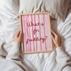 Op de afbeelding: Een ingelijst kunstwerk met een licht houten frame. Het kunstwerk toont verticale roze en witte strepen met de tekst "What's for pudding?" in een handschriftstijl. Het kunstwerk wordt vastgehouden door een persoon die een witte trui draagt.