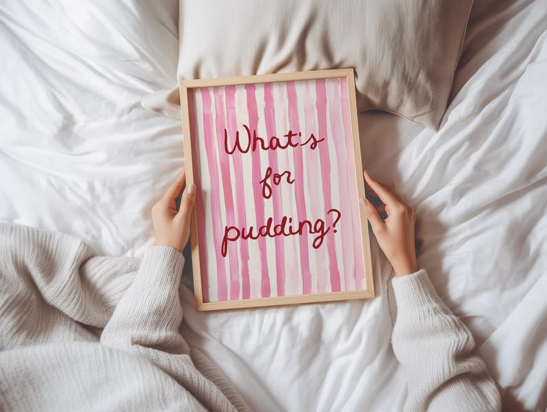 Op de afbeelding: Een ingelijst kunstwerk met een licht houten frame. Het kunstwerk toont verticale roze en witte strepen met de tekst "What's for pudding?" in een handschriftstijl. Het kunstwerk wordt vastgehouden door een persoon die een witte trui draagt.