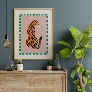 May include: A framed print of a cheetah sitting on a pink background with a green and white checkered border. The cheetah is facing away from the viewer and has a black tail.