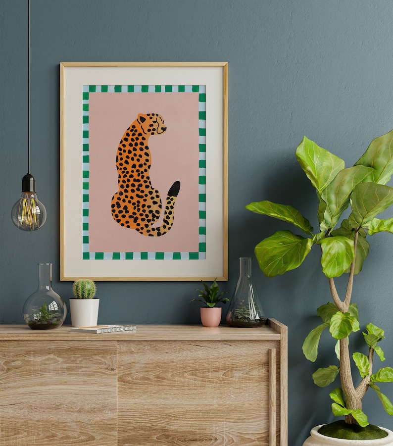 May include: A framed print of a cheetah sitting on a pink background with a green and white checkered border. The cheetah is facing away from the viewer and has a black tail.