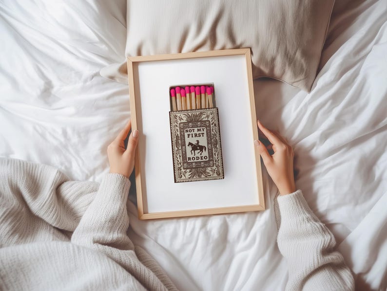 May include: A rectangular wooden frame containing a print of a matchbox with bright pink tips. The matchbox features a vintage-style illustration of a cowboy on a horse with the text "NOT MY FIRST RODEO" printed in black on a cream background.
