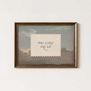 K&ouml;nnte beinhalten: Gerahmtes Kunstwerk mit einer Landschaftsmalerei mit bew&ouml;lktem Himmel. Ein cremefarbenes Papier mit gezackten Kanten und dem Text "HOW LUCKY ARE WE" ist auf das Gem&auml;lde gelegt. Der Rahmen ist dunkel und aufwendig goldfarben.