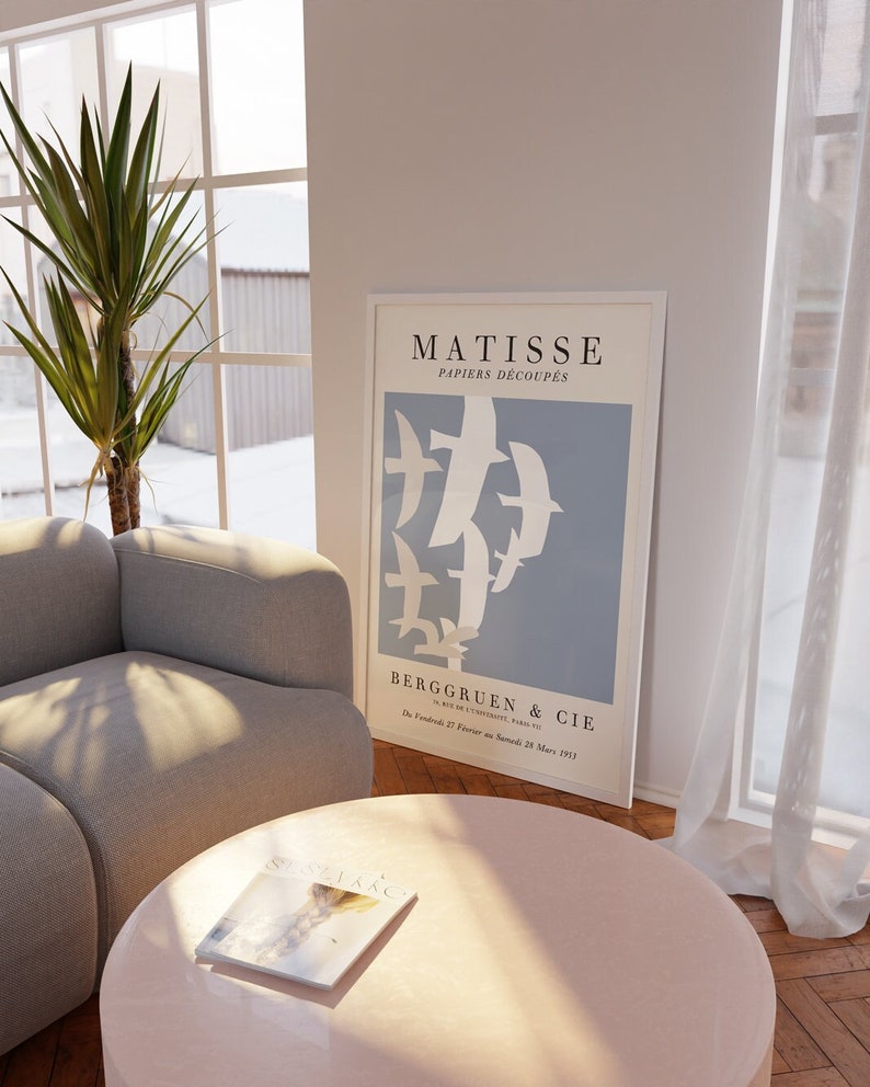 May include: A framed poster with a blue background and white paper cutouts of birds. The poster is titled "Matisse Papiers Decoupes" and has the text "Berggruen & Cie" at the bottom. The poster is hanging on a white wall in a living room with a couch and a coffee table.