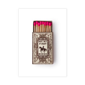 May include: A rectangular matchbox containing wooden matches with bright neon pink tips. The box features a brown and white decorative border with a central illustration of a person on a horse and the text NOT MY FIRST RODEO printed in black.