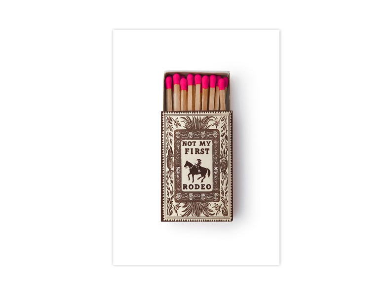 May include: A rectangular matchbox containing wooden matches with bright neon pink tips. The box features a brown and white decorative border with a central illustration of a person on a horse and the text NOT MY FIRST RODEO printed in black.
