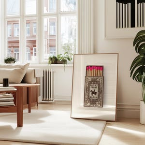 May include: A framed wall art print featuring a vintage-style matchbox illustration with the text NOT MY FIRST RODEO and a graphic of a cowboy on a horse. The matchbox is open, revealing a row of wooden matches with bright pink tips.