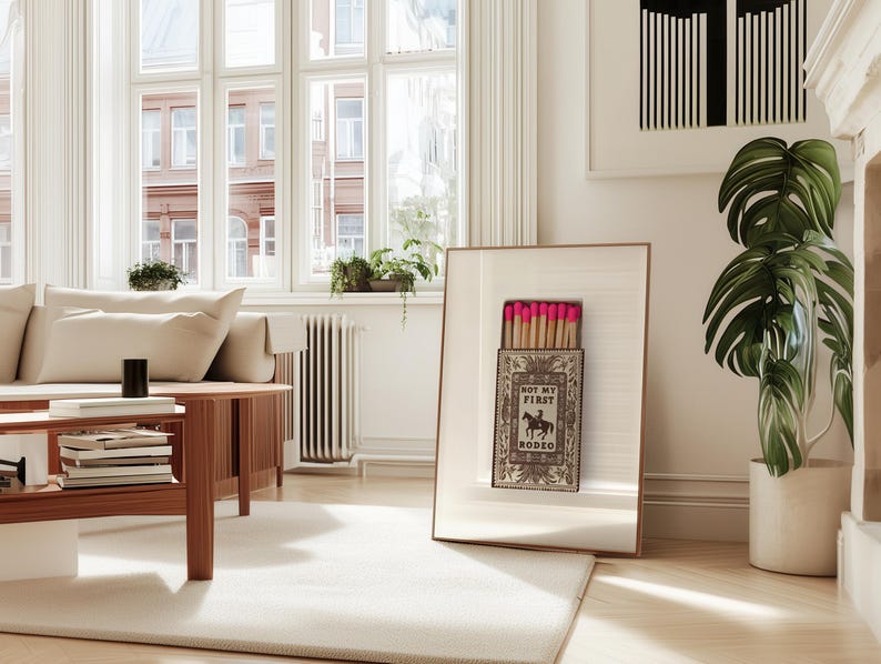 May include: A framed wall art print featuring a vintage-style matchbox illustration with the text NOT MY FIRST RODEO and a graphic of a cowboy on a horse. The matchbox is open, revealing a row of wooden matches with bright pink tips.