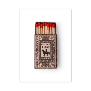 May include: A rectangular matchbox with a light brown and dark brown decorative border featuring floral motifs. The center displays the text "NOT MY FIRST RODEO" above a silhouette of a person riding a horse. The box contains wooden matches with red tips.