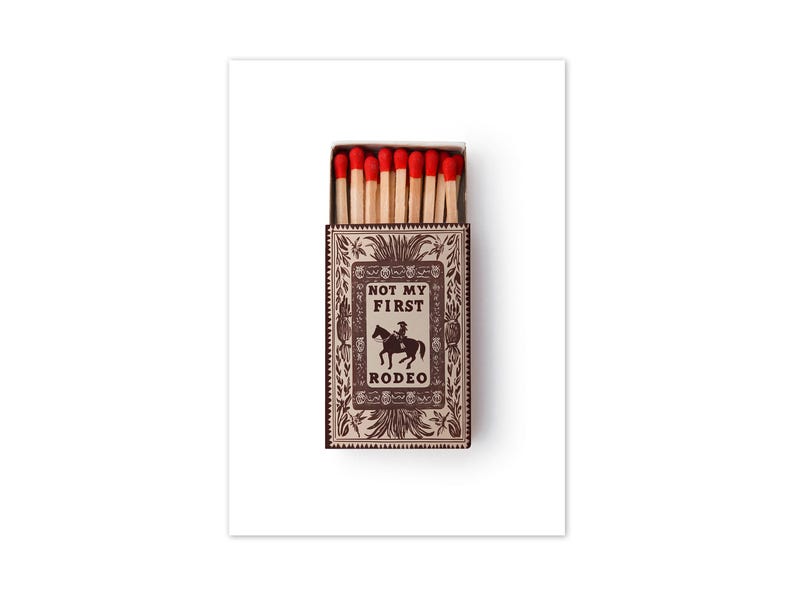 May include: A rectangular matchbox with a light brown and dark brown decorative border featuring floral motifs. The center displays the text "NOT MY FIRST RODEO" above a silhouette of a person riding a horse. The box contains wooden matches with red tips.