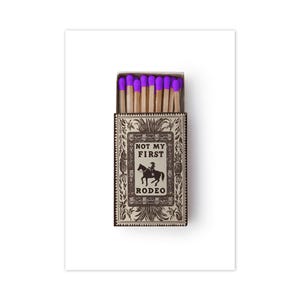 May include: A rectangular matchbox containing wooden matches with bright purple tips. The box features a tan and dark brown decorative border with a central illustration of a person riding a horse and the text "NOT MY FIRST RODEO" printed in black.
