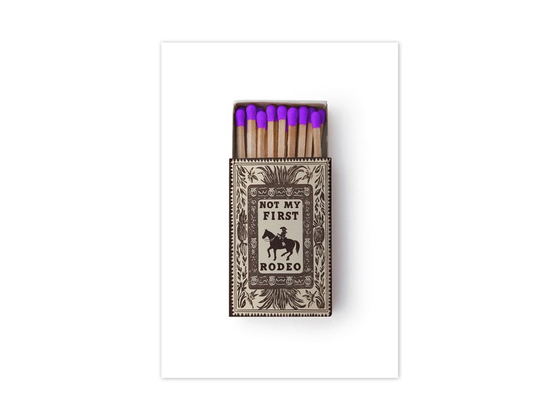 May include: A rectangular matchbox containing wooden matches with bright purple tips. The box features a tan and dark brown decorative border with a central illustration of a person riding a horse and the text "NOT MY FIRST RODEO" printed in black.