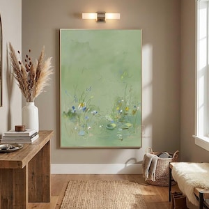 May include: An abstract painting with a light green background and a gold frame. The artwork depicts a pond scene with lily pads and flowers in shades of blue, yellow, and white. The painting hangs above a wooden table and a woven basket.