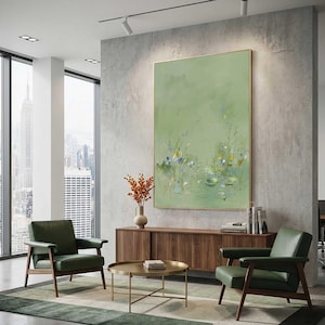 May include: A large, framed abstract painting with a light green background and delicate floral details. The artwork hangs above a wooden cabinet, complemented by two green armchairs and a gold coffee table. The room has large windows overlooking a cityscape.