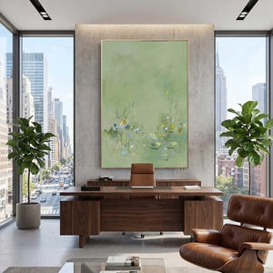 May include: An abstract painting with a light green background and a gold frame. The artwork depicts a pond scene with water lilies and reeds. The painting hangs above a wooden desk in a modern office setting.