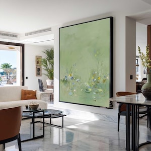 May include: Large abstract painting with a black frame. The artwork features a light green background with delicate floral and aquatic details in shades of blue, yellow, and white. The painting hangs on a white wall in a modern living space.
