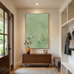 May include: A modern entryway featuring a large abstract painting in shades of green, brown, and blue. A wooden cabinet with a vase of dried flowers and a gold geometric sculpture sits below the painting. The room has a neutral color palette.