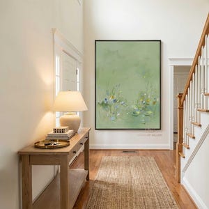 May include: An abstract painting in a black frame hangs on a wall in a hallway. The painting features a light green background with delicate floral and aquatic details. The hallway has a wooden console table, a lamp, and a woven rug.