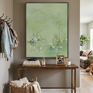 May include: A large framed abstract painting in shades of green, depicting a pond scene with lily pads and flowers. A wooden console table displays books, a photo frame, and a candle. A woven basket with a blanket sits below.
