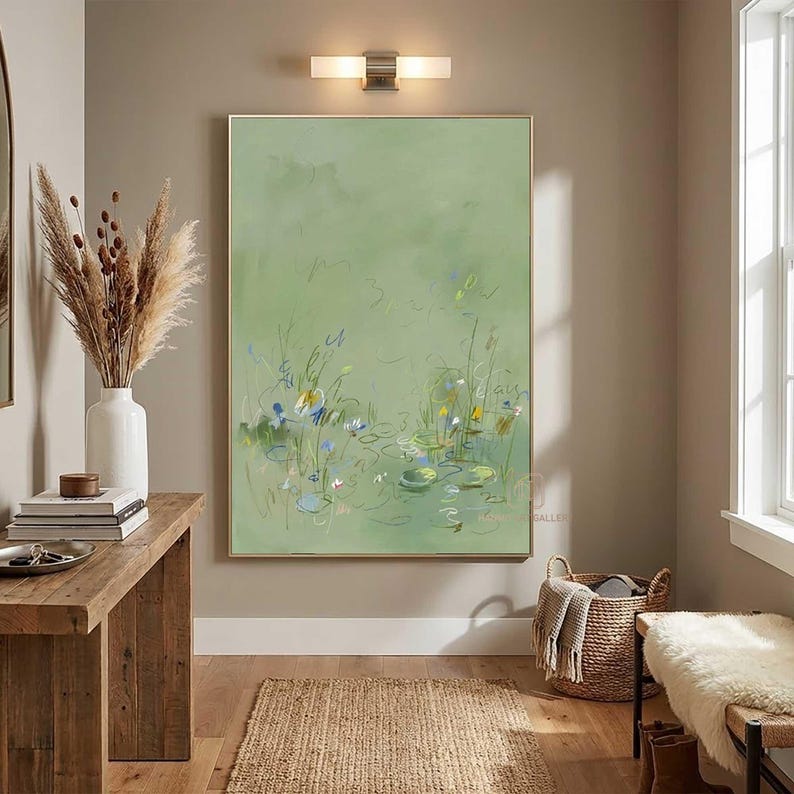 May include: An abstract painting with a light green background and a gold frame. The artwork depicts a pond scene with lily pads and flowers in shades of blue, yellow, and white. The painting hangs above a wooden table and a woven basket.